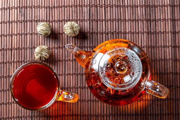 Tea in a glass teapot with a blooming large flower. Teapot with exotic green tea-balls blooms flower and a glass Cup of tea. Tea ceremony on a dark background