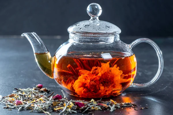 Tea in a glass teapot with a blooming large flower. Teapot with exotic green tea-balls blooms flower and dried tea and rose buds