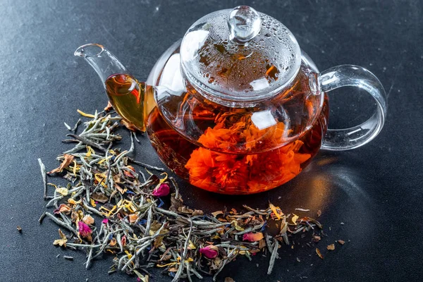 Tea in a glass teapot with a blooming large flower. Teapot with exotic green tea-balls blooms flower and dried tea and rose buds