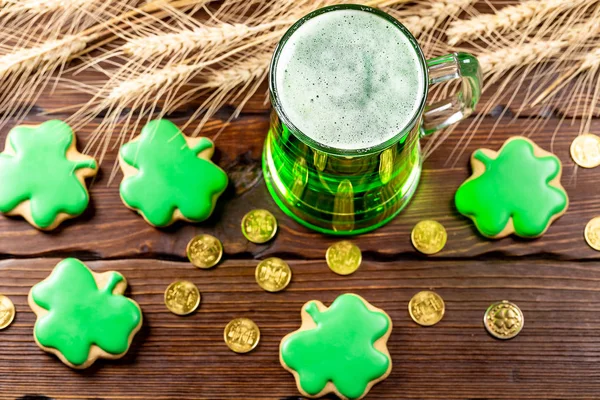 Green beer in a glass mug with gingerbread clover, horseshoe, wheat spike and gold coins on a rustic wooden surface. Festive background for St. Patrick\'s day.