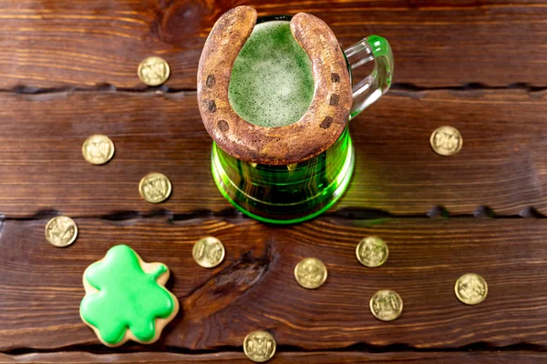 Green beer in a glass mug with gingerbread clover, horseshoe and gold coins on a rustic wooden surface. Festive background for St. Patrick's day.