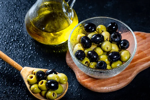 Aceite de oliva en botellas de vidrio vintage con aceitunas negras, verdes y cuchara de madera sobre fondo negro. Copyspace. Flatley. Fondo alimentario con gotas de agua.Comida saludable y concepto de dieta — Foto de Stock