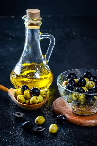 Aceite de oliva en botellas de vidrio vintage con aceitunas negras, verdes y cuchara de madera sobre fondo negro. Copyspace. Flatley. Fondo alimentario con gotas de agua.Comida saludable y concepto de dieta — Foto de Stock