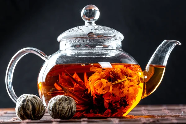 Tea in a glass teapot with a blooming large flower. Teapot with exotic green tea-balls blooms flower. Tea ceremony on a dark background