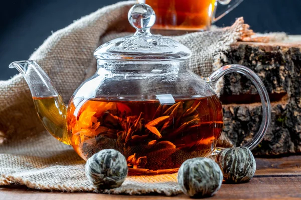 Tea in a glass teapot with a blooming large flower. Teapot with exotic green tea-balls blooms flower and a glass Cup of tea. Tea ceremony on the background of burlap and old wooden stump
