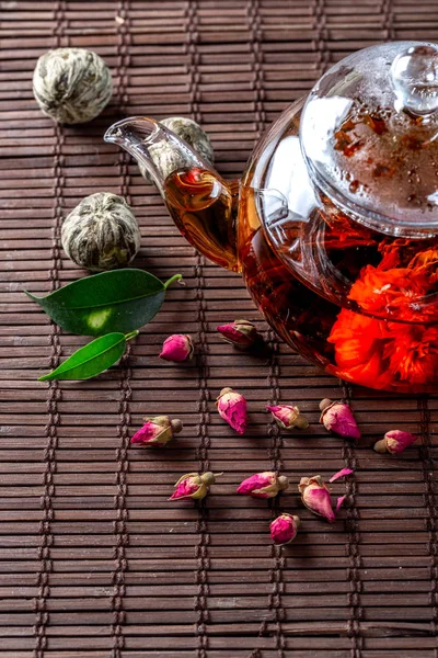 Tea in a glass teapot with a blooming large flower. Teapot with exotic green tea-balls blooms flower and dried rose buds with fresh tea leaves and dry.