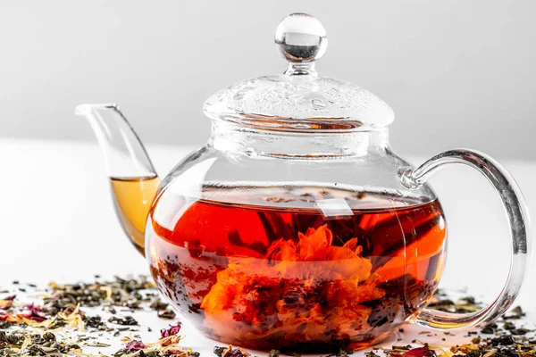 Tea in a glass teapot with a blooming large flower. Teapot with exotic green tea on a white background with scattered dried tea with petals and pieces of fruit — Stock Photo, Image