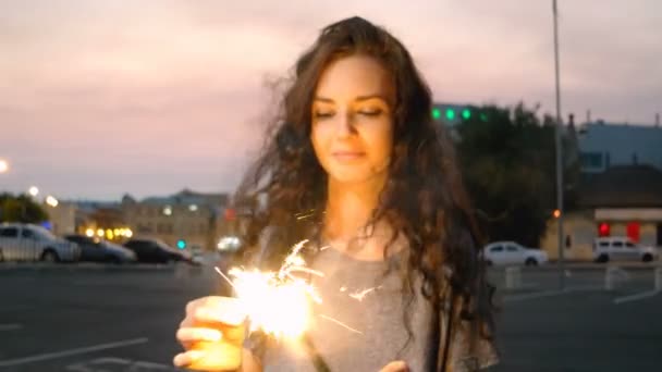 Menina sorridente começar sparkler por jogo na rua da noite com luzes da cidade fora de foco no fundo . — Vídeo de Stock
