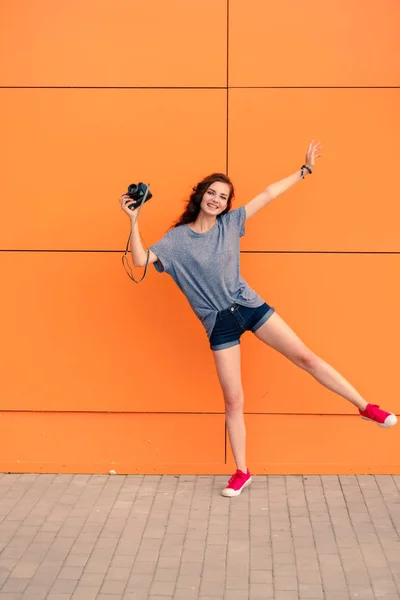 Gadis cantik berpose di satu kaki dengan kamera vintage di depan dinding oranye — Stok Foto