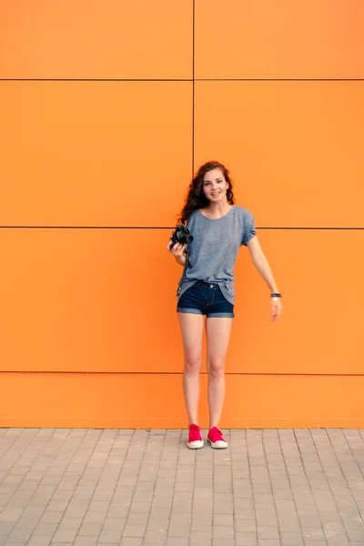 Salido chica Hipster con cámara de fotos vintage en una mano de pie delante de la pared naranja, espacio de copia, imagen tonificada — Foto de Stock