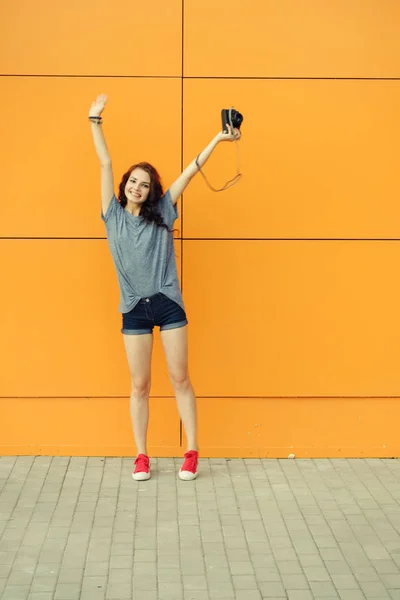 Hipster κορίτσι με vintage φωτογραφία κάμερα σε ένα χέρι αυξήσει τα χέρια της στο μέτωπο του πορτοκαλί τοίχο, αντιγράψτε χώρο, τονωμένο εικόνα — Φωτογραφία Αρχείου
