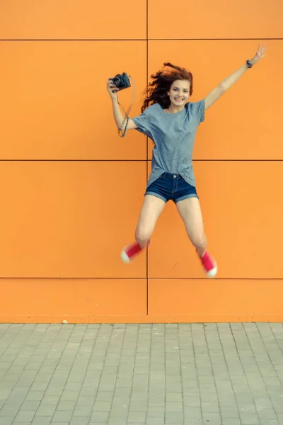 Pretty girl jamping with vintage camera in front of orange wall — Stock Photo, Image