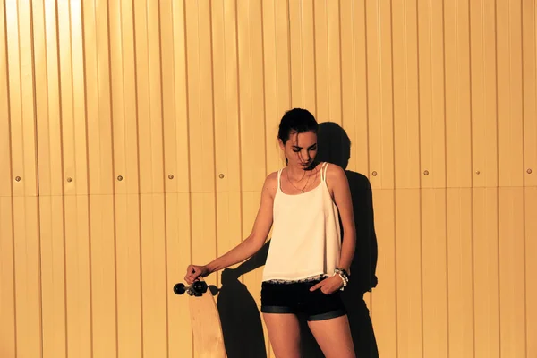 Menina em shorts jeans e top tanque de pé com skate na frente da cerca mwtal amarelo e olhando para baixo — Fotografia de Stock