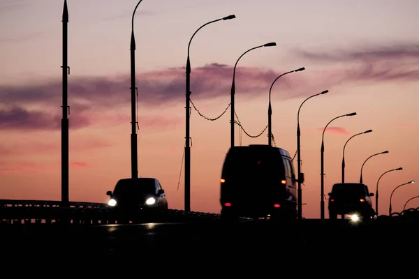 Siluetas de coches y furgonetas delante del cielo del atardecer con siluetas de farolas — Foto de Stock