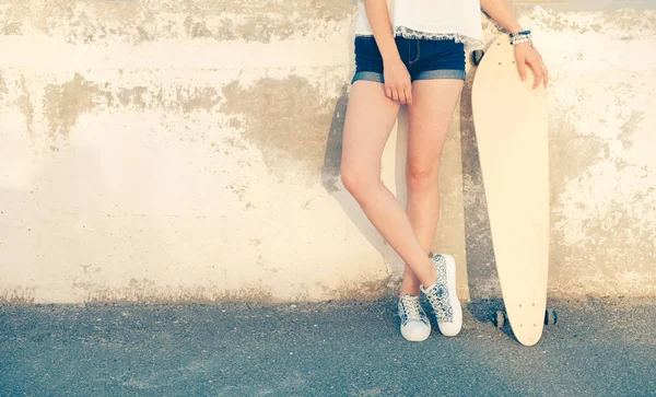 Mädchen, das in der Nähe der Wand steht, die Beine gekreuzt und ihr Longboard zurückgelehnt hält, verschlissene Wand, viel Kopierraum — Stockfoto