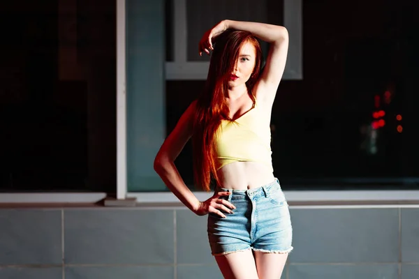 Ginger haired girl, portrait in night city setting. Street fashion style portrait of young pretty woman with long red hair weared denim shorts and yellow tanktop posing half face hidden by hair