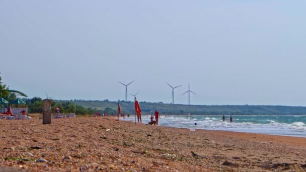 Les éoliennes tournent en arrière-plan pendant que les gens s'amusent sur la plage au premier plan — Video