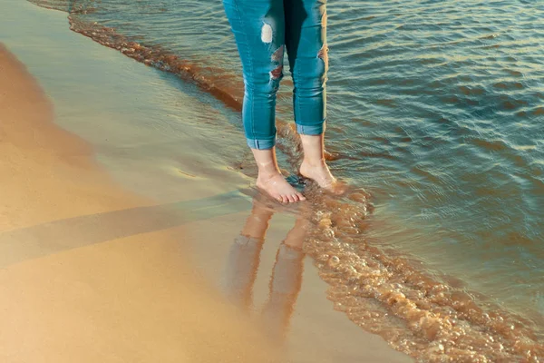 Gambe di ragazza a piedi nudi in jeans strappati sul bordo dell'acqua sulla spiaggia nel tempo del tramonto — Foto Stock