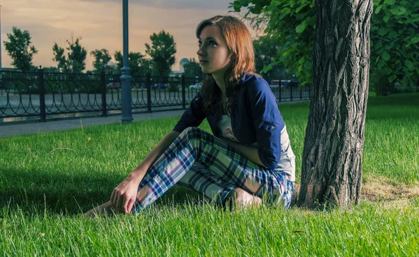 自由奔放に生きる若い赤毛の女の子草バックライトの上に座ってとよそ見。カラフルな夕日の光. — ストック写真