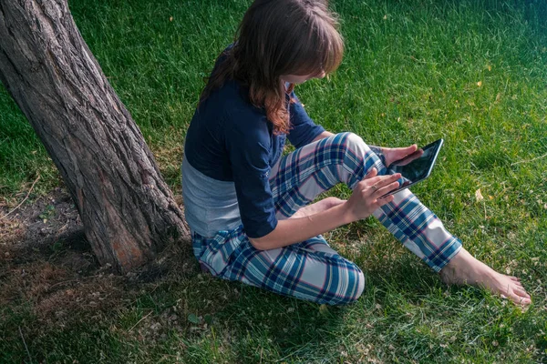 Unga rödhårig kvinna i chekered slitage med en tablet-pc i parken lutad mot ett gammalt träd som hon rullar använda pekskärmen — Stockfoto