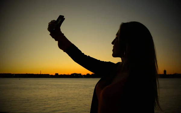 Sylwetka młoda kobieta biorąc selfie naprzeciwko rzeki zachód słońca — Zdjęcie stockowe