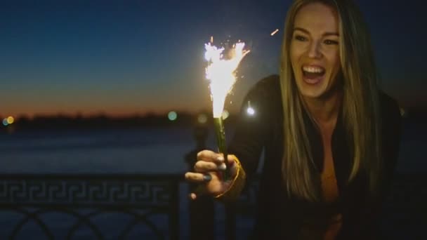 Young happy woman stands on riverside with sparkler in front of sunset light. — Stock Video