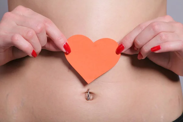 善良的女人在她扁平的肚子上握着一颗红色的纸心 肚脐刺痛 怀孕后皮肤伸展的最初迹象可见于她光滑的胃部 — 图库照片