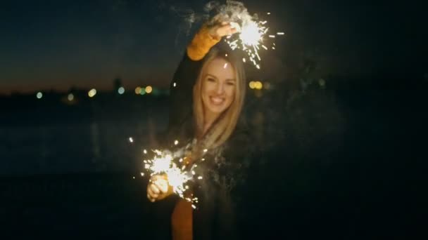 Mujer alegre sosteniendo dos bengalas ardientes en las manos y bailando al aire libre. Chica rubia celebrando la víspera de año nuevo con luces de bengala. Hermosa mujer sosteniendo un chispeante palos en slomo noche de fiesta. — Vídeo de stock