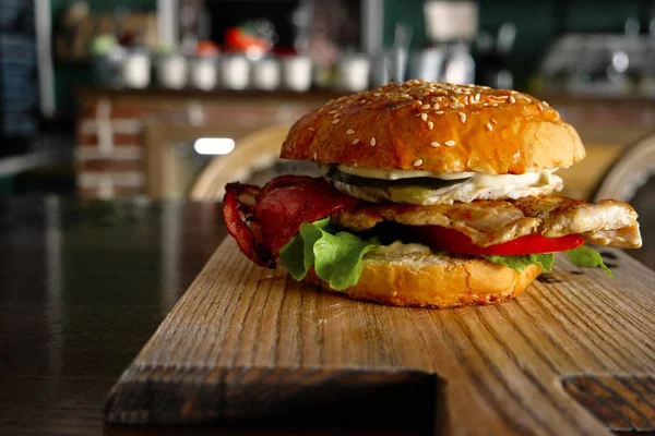 Burger s rozřezanou vajíčkem a backon servírovaný na řezací desce s rozmazanou interiérem na pozadí — Stock fotografie