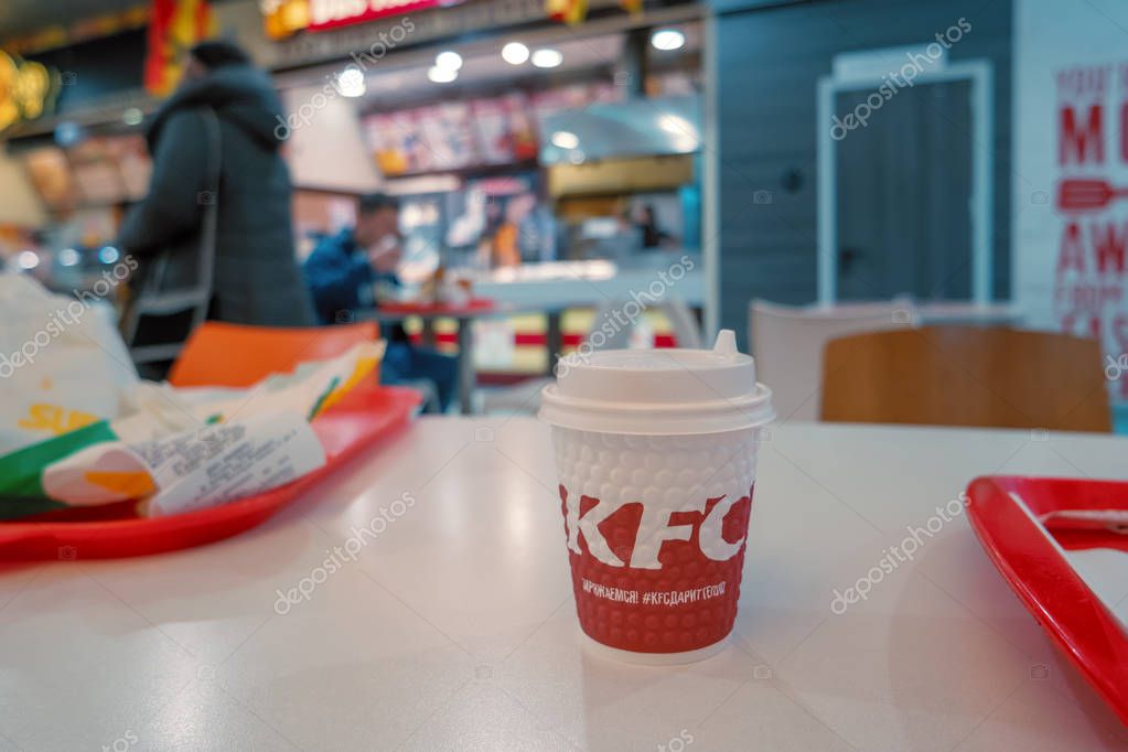 Astrakhan, Russia - 13 of Feb. 2019: Takeaway coffee cup with KFC logo on the side standing on table of shopping mall food court with eating unrecognizable people on background