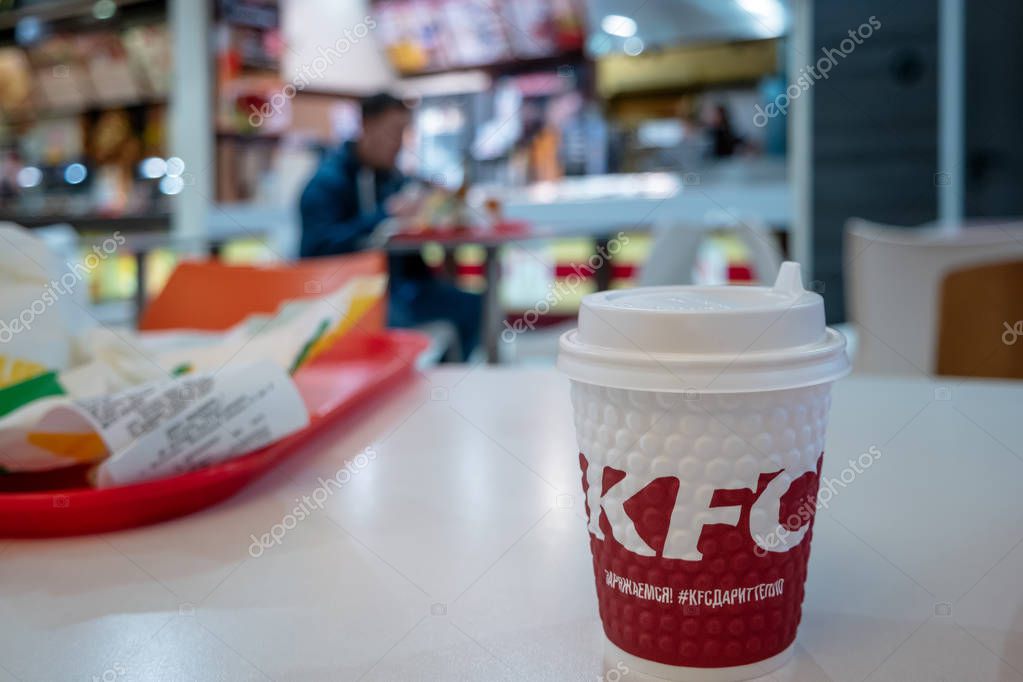 Astrakhan, Russia - 13 of Feb. 2019: Take-away coffee cup with KFC fast food brand logo on the side standing on table of shopping mall food court with eating unrecognizable people on background