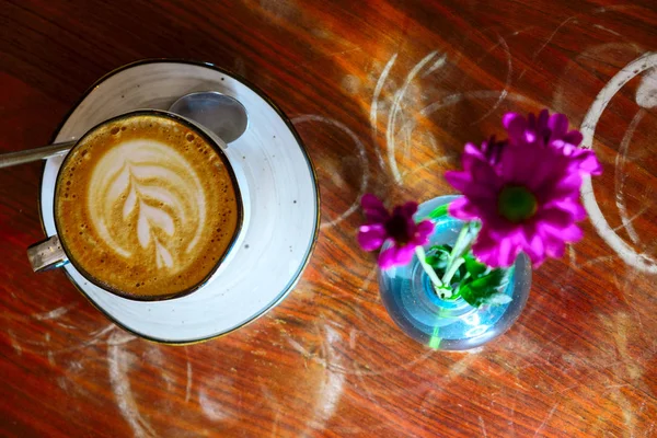 Šálek kávy s kapučíkem a květinami v láhvi na opotřebené naleštěné stolní desce — Stock fotografie