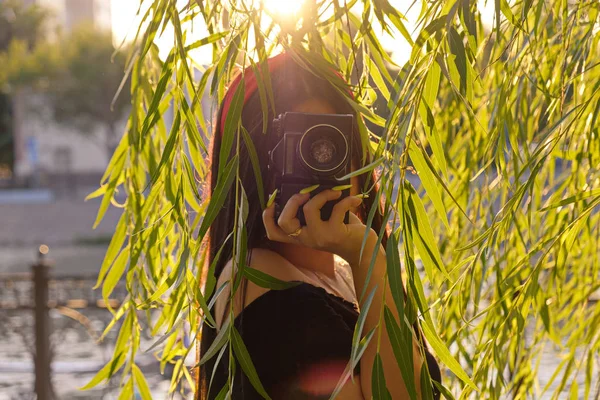 在日落时间，在公园与相机的亚洲女孩。布鲁内特女孩与电影相机在柳树前的复古彩色图像 — 图库照片