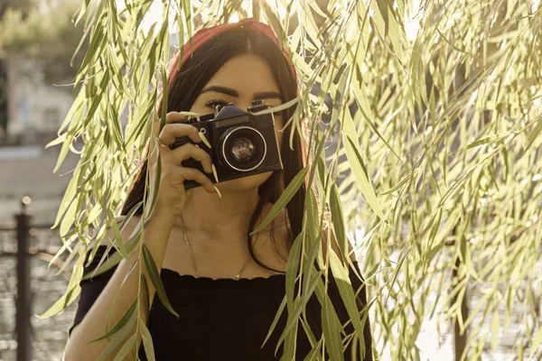 Menina asiática com câmera no parque na hora do pôr do sol. Menina morena com câmera de filme na imagem colorida vintage escondendo no salgueiro — Fotografia de Stock