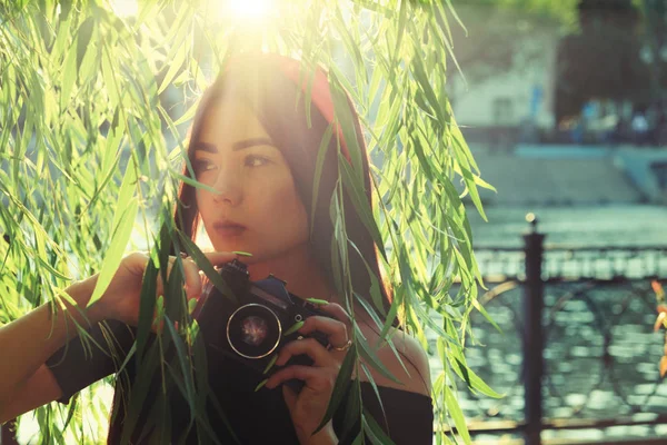 夕日の時間に公園にカメラを持つアジアの女の子。柳の枝に対してポーズヴィンテージ色の画像でフィルムカメラを持つブルネットの女の子 — ストック写真