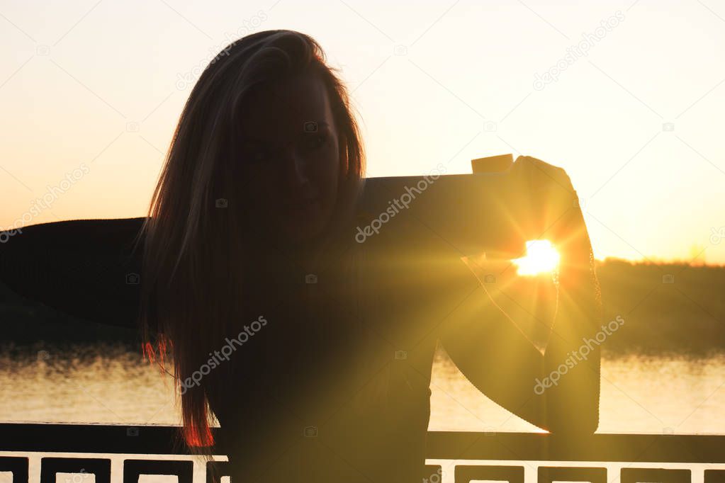 Adult blond hair woman with scateboard on her shoulders posing outside on waterfront in front of sunset