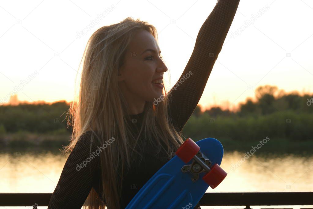 Blond hair scateboarder holds her scateboard by hand and greets somebody by another in front of sunset on waterfront
