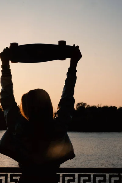 Donna posa con le mani tenendo lo skateboard sollevato nel cielo del tramonto — Foto Stock