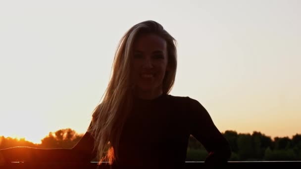 Mujer rubia adulta de unos 30 años se para en el paseo marítimo con el pelo iluminado por el sol al atardecer y después de dar la vuelta. Slomo retroiluminado . — Vídeo de stock