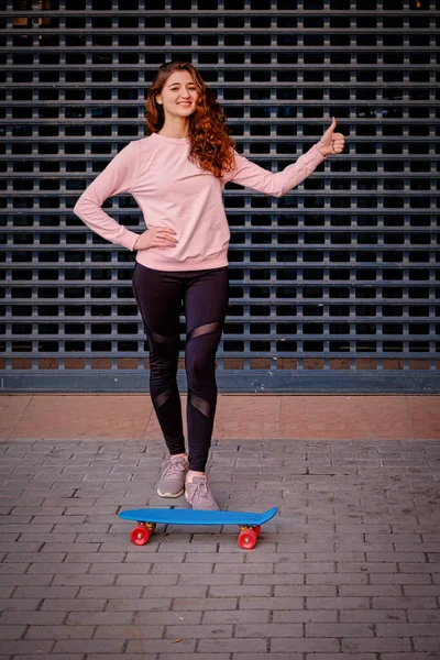 Спортивная девушка стоит рядом со своим скейтбордом и показывает нам большой палец вверх знак — стоковое фото