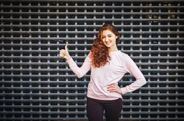 Joyful redhaired girl shows thumb-up sign of appruval. Model is posing in front of metal grid.