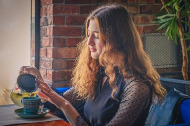 Kızıl saçlı kadın önündeki masanın üstündeki fincana bitki çayı döküyor..