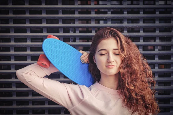 Ginger haired lady daydreaming with her eyes closed. She is holding her blue pennyboard on her shoulder