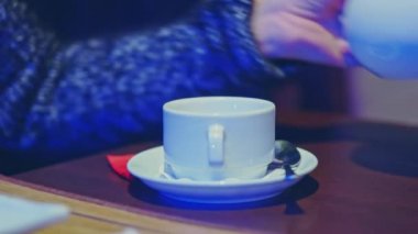 Bir kadın kafede bir masaya oturur ve neon ışığındaki çaydanlıktan kendine bitki çayı doldurur.