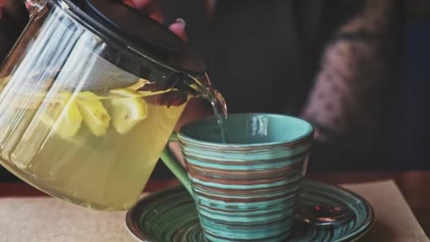 Lass versando tisana calda da un bollitore di vetro trasparente dove le erbe al vapore, menta e alcune fette di limone galleggiano in una tazza di ceramica a strisce. — Video Stock