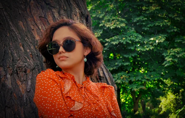 Chica asiática posa en gafas de sol retro apoyadas en el tronco de un enorme árbol en el bosque. Ella está vestida con un vestido vintage de color rojo y a la derecha hay mucho espacio para el texto. —  Fotos de Stock