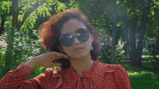 Lovely Asian woman with short curvy hair poses in the park dressed in a retro dress and sunglasses — Stock Video