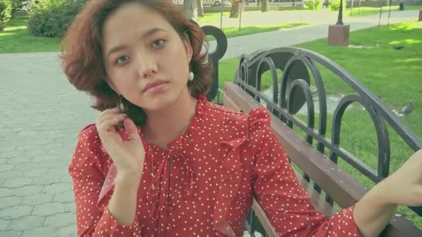 Asian lady sitting on bench looking at camera smiling. Pretty young multicultural woman enjoying summer in park on park bench — Stock Video