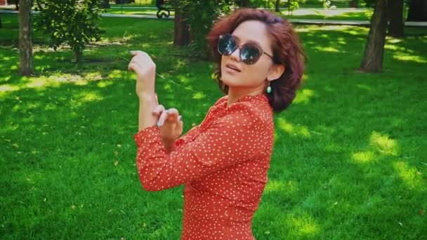 Schöne Koreanerin in Retro-Sonnenbrille und rotem Kleid tanzt auf grünem Gras im Park, Slomo — Stockvideo