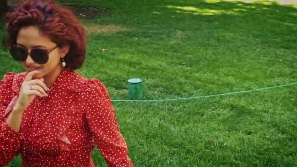 Bonita mujer asiática en gafas de sol retro y vestido rojo bailando sobre hierba verde en parque — Vídeo de stock